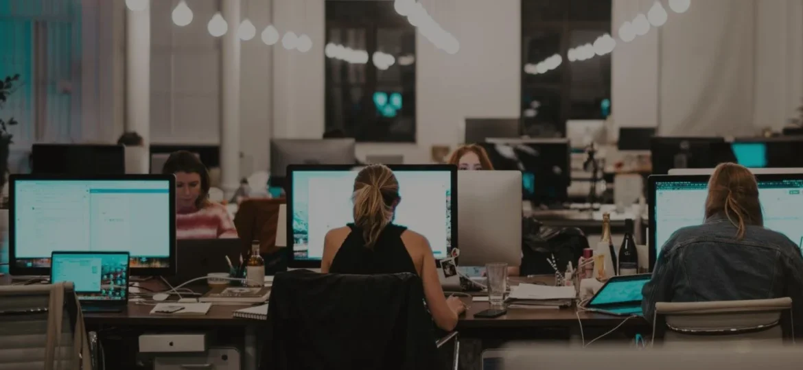 Office scene captured in a lower-light setting, showing various employees engaged at their workstations, highlighting a productive and focused atmosphere.